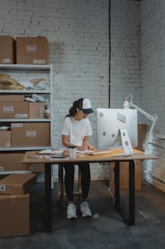pexels photo 6170176 6170176 Woman in a warehouse setting organizing packages on a desk, showcasing logistics and office work.